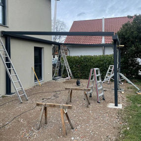 Montage einer Terrassenüberdachung an einem modernen Wohnhaus: bereits montierte Aluminiumträger an der Fassade und auf vorderen Pfosten, mehrere Leitern und Montageböcke auf vorbereiteter Erdfläche, Werkzeuge und Kabel verteilt im Arbeitsbereich, Hecke und Nachbarhaus im Hintergrund – Dokumentation eines Installationsschritts für Überdachungssysteme von ML DER RAUM.