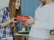 A woman gives a man a red gift in a cozy kitchen. Both are smiling. She wears a plaid shirt; he wears a white top. Blue dishes visible.