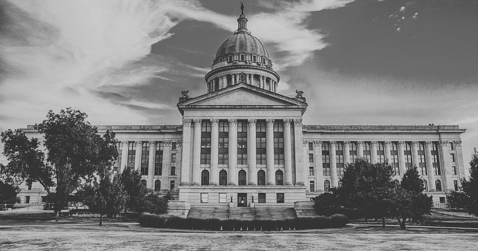 Black-and-white photo of the Oklahoma State Capitol with a central dome, columns, trees, and clouds; STATE OF OKLAHOMA visible.