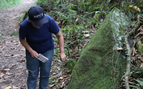 Uma mulher está ao ar livre em um caminho de terra cercado por folhagens. Ela está vestindo uma camisa azul escura, calça jeans azul e um boné preto com a palavra "Dakila" escrita. Ela estão segurando um caderno, na mão esquerda. Seu braço direito é estendido para baixo, apontando para uma grande rocha coberta de musgo que está situada à sua frente com marcas horizontais com aspectos não naturais.