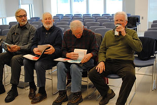 Four men sitting in classroom chairs chat and look through music while they wait for a singing club to start.