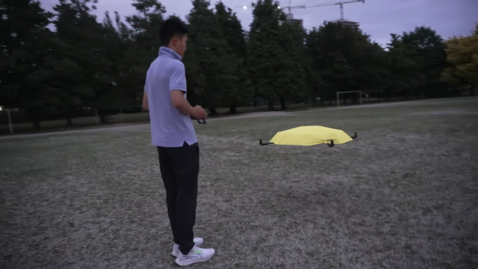 Man in blue shirt flying a yellow drone in a park at dusk. Trees and construction cranes are visible in the background.