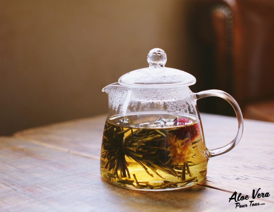 Théière en verre sur table en bois, infusion dorée avec fleurs et feuilles, lumière chaude, logo Aloe Vera Pour Tous.