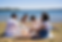 Four friends enjoy a picnic by a lake, sitting on a blanket. They're smiling, surrounded by food baskets. The sky is clear and sunny.