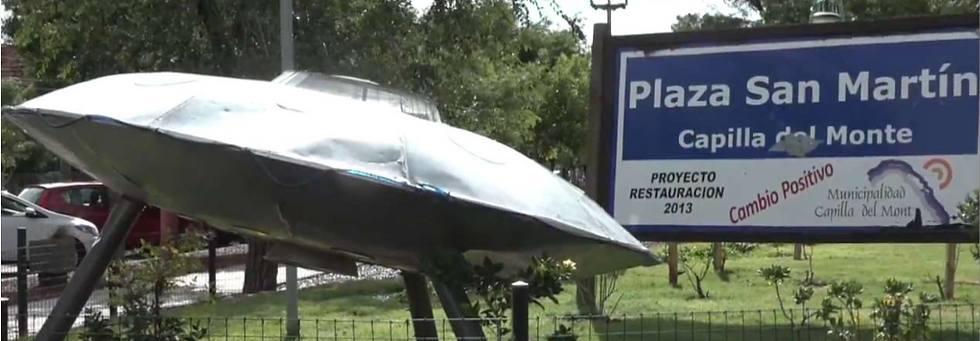 Wide landscape photo of a metallic UFO sculpture in a park next to a blue sign reading Plaza San Martín, Capilla del Monte.