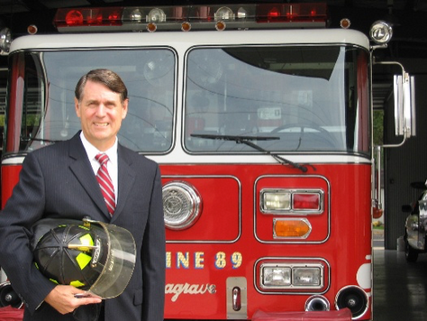 Man in suit in front of a fire truck holding firefighter helmet.