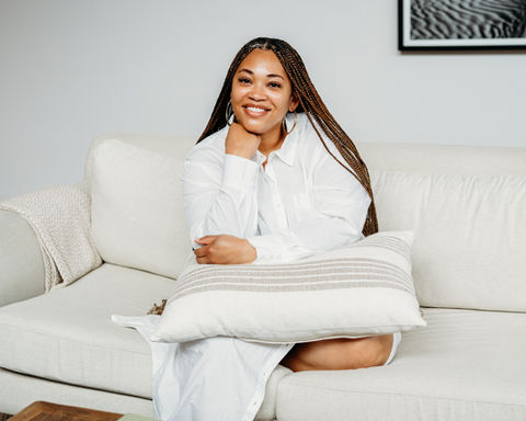 Smiling woman sitting on white couch holding a pillow, looking at camera.