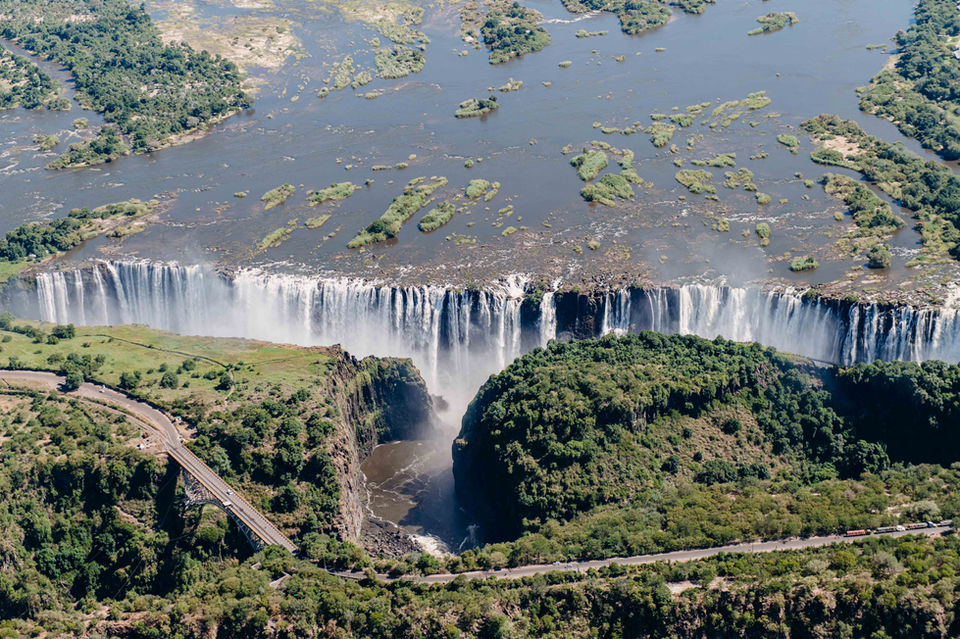 • Aerial view of Victoria Falls from a helicopter.
• Helicopter flying above Victoria Falls and the Zambezi River.
• Wide aerial shot showing the full span of Victoria Falls.
• Close-up photo of water cascading over Victoria Falls.
• Trail walkway leading to viewpoints at Victoria Falls.
• Mist rising from Victoria Falls during the daytime.
• View of the falls with water crashing into the gorge below.
• Overlook point showing the width and depth of Victoria Falls.
• A visitor is standing near the edge of Victoria Falls viewpoint.
• Thunderous water flow captured along the trail.
• The Zambezi River is flowing toward the falls.
• The picturesque scenery envelops Victoria Falls.
• Family photo at a Victoria Falls lookout point.
• A rainbow is forming in the mist above Victoria Falls.
• Pathway through the Victoria Falls rainforest area.