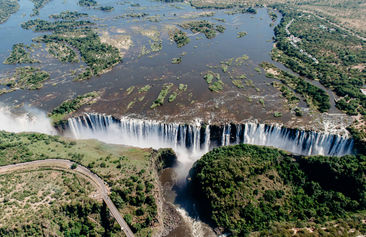 • Aerial view of Victoria Falls from a helicopter.
• Helicopter flying above Victoria Falls and the Zambezi River.
• Wide aerial shot showing the full span of Victoria Falls.
• Close-up photo of water cascading over Victoria Falls.
• Trail walkway leading to viewpoints at Victoria Falls.
• Mist rising from Victoria Falls during the daytime.
• View of the falls with water crashing into the gorge below.
• Overlook point showing the width and depth of Victoria Falls.
• A visitor is standing near the edge of Victoria Falls viewpoint.
• Thunderous water flow captured along the trail.
• The Zambezi River is flowing toward the falls.
• The picturesque scenery envelops Victoria Falls.
• Family photo at a Victoria Falls lookout point.
• A rainbow is forming in the mist above Victoria Falls.
• Pathway through the Victoria Falls rainforest area.