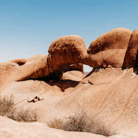 Person relaxing under an arch-shaped rock formation in the desert landscape.