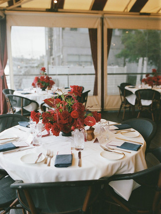 Round table set with white table cloth and red floral arrangement.
