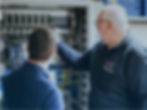 Zwei SPS-Techniker untersuchen komplexe Verkabelungen in einem offenen Serverschrank. Einer zeigt auf die Komponenten und deutet damit an, dass er aktiv Problemlösungs- oder Schulungsaufgaben übernimmt.