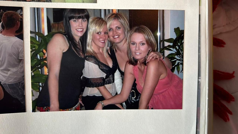 Four women pose closely together, smiling in a warmly lit room with green plants. The mood is cheerful, creating a sense of friendship.