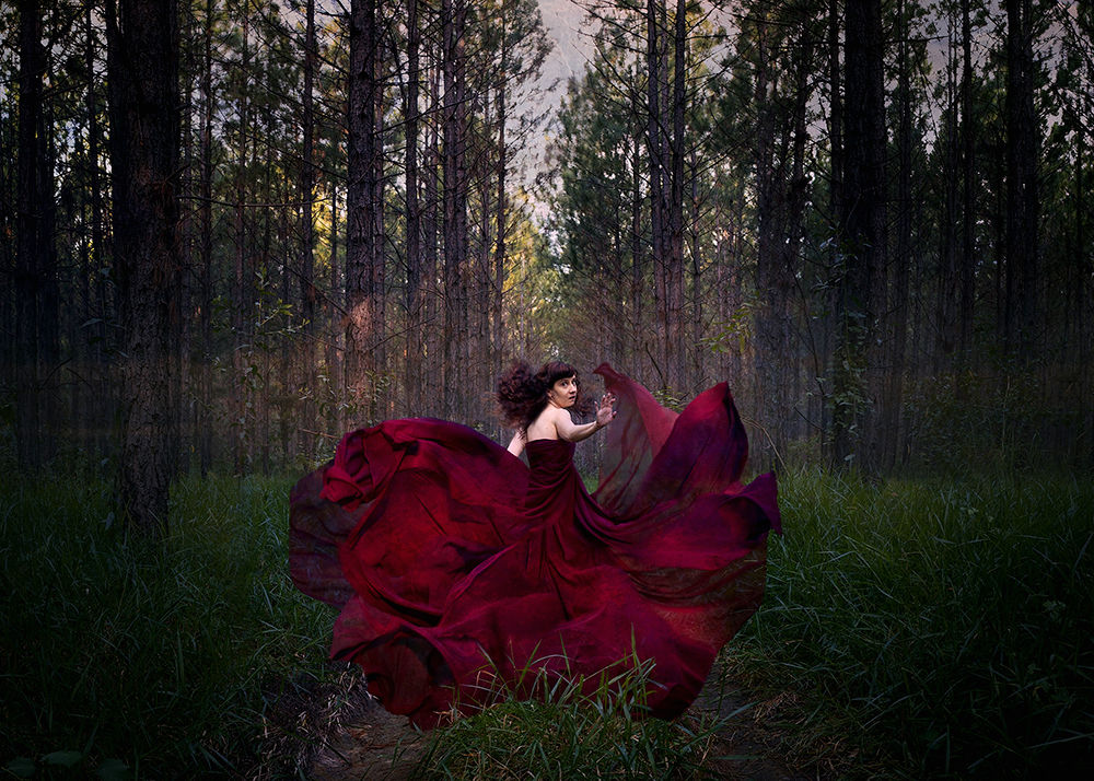 A conceptual fine art photo of a girl in a flowing red dress running through a forest