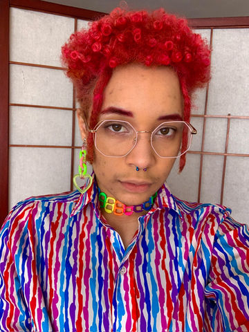 Keiko is a Black nonbinary artist with short pink hair, colorful septum ring,
and pastel pink glasses pictured with a neutral expression. They're wearing a
white and mixed berry striped button down and their own handmade
jewelry: a neon green heart earring and a neon rainbow pull tab choker.