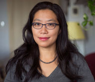 Haisi Hu is an Asian woman. She has long hair to her shoulder. She is sitting
in a living room next to some plants and lamp. She wears a grey sweater,
red lipstick and a grey pearl necklace. She smiles and looks straight to the
camera. She is in her mid forties.