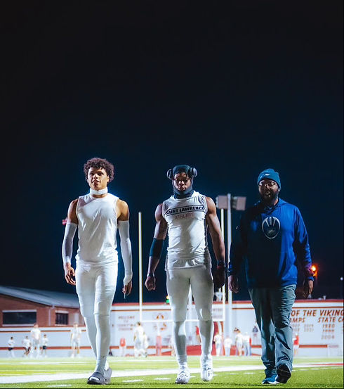 Three men are walking side by side on a football field at night. The two men on the left are wearing white athletic gear, with the middle one wearing a sleeveless shirt that says "Saint Lawrence Athlete." The man on the right is dressed in casual clothes, wearing a blue jacket, gray pants, and a blue beanie. The background shows a stadium with bright lights and a few people in white uniforms.