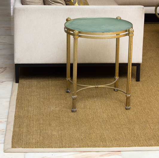 Round glass and gold metal side table sitting on a bordered tan woven rug next to a modern beige sofa.