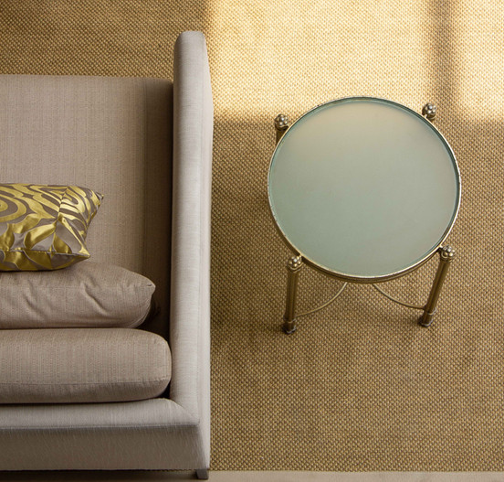 Top-down view of a glass side table, the armrest of a beige sofa with two pillows, and a textured tan area rug.