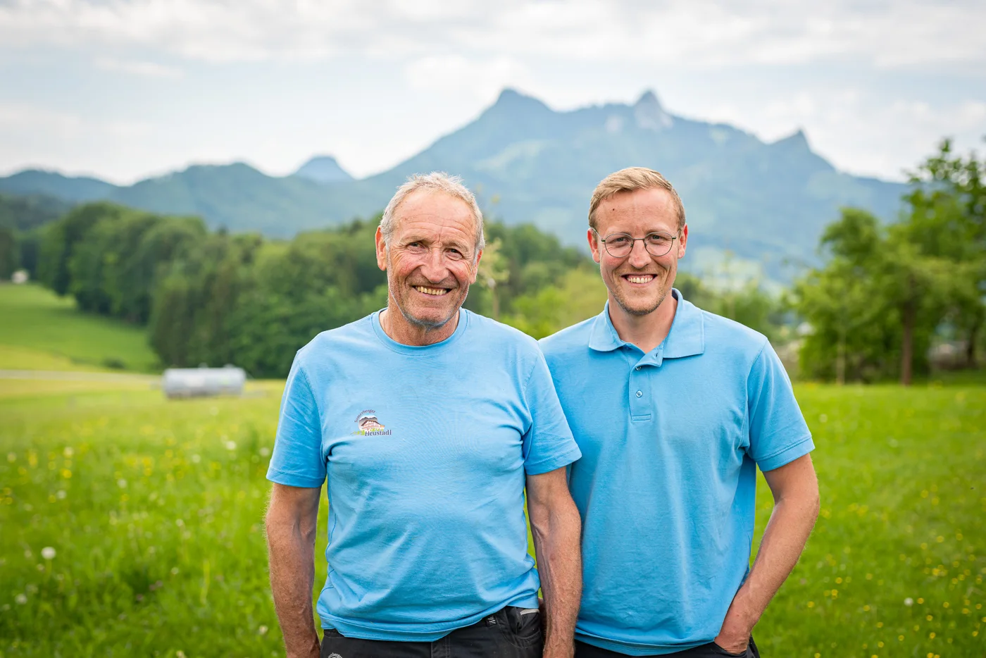 Zwei Männer in hellblauen Shirts auf einer Wiese vor Bergkulisse