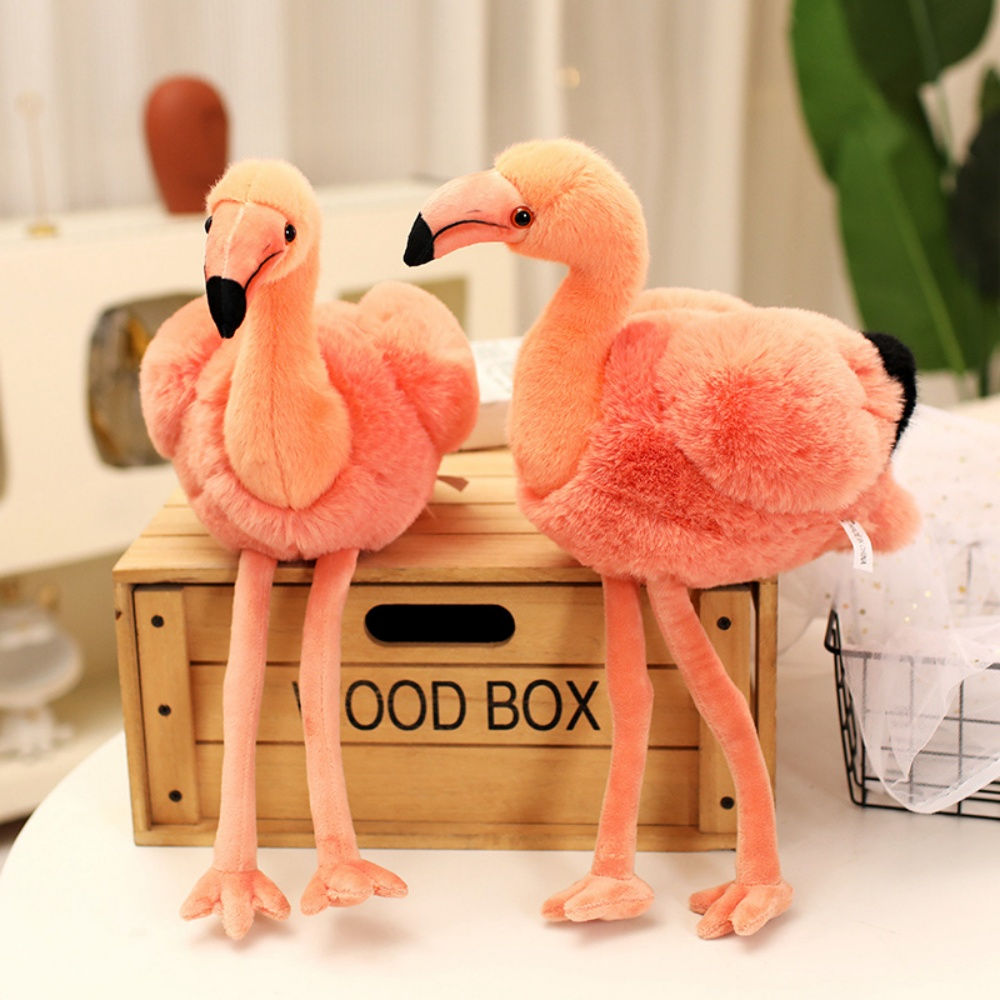 Two fluffy pink flamingo plush toys sitting side-by-side on a wooden crate, with their long legs hanging over the edge.