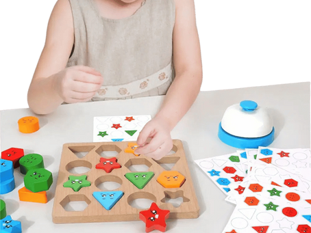 Young girl sorts colorful wooden shapes into a puzzle board at a table, with pattern cards and a bell beside her.