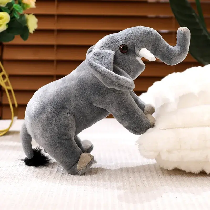 A cute grey elephant plush toy with a raised trunk and white tusks, resting its front legs on a fluffy white cushion.