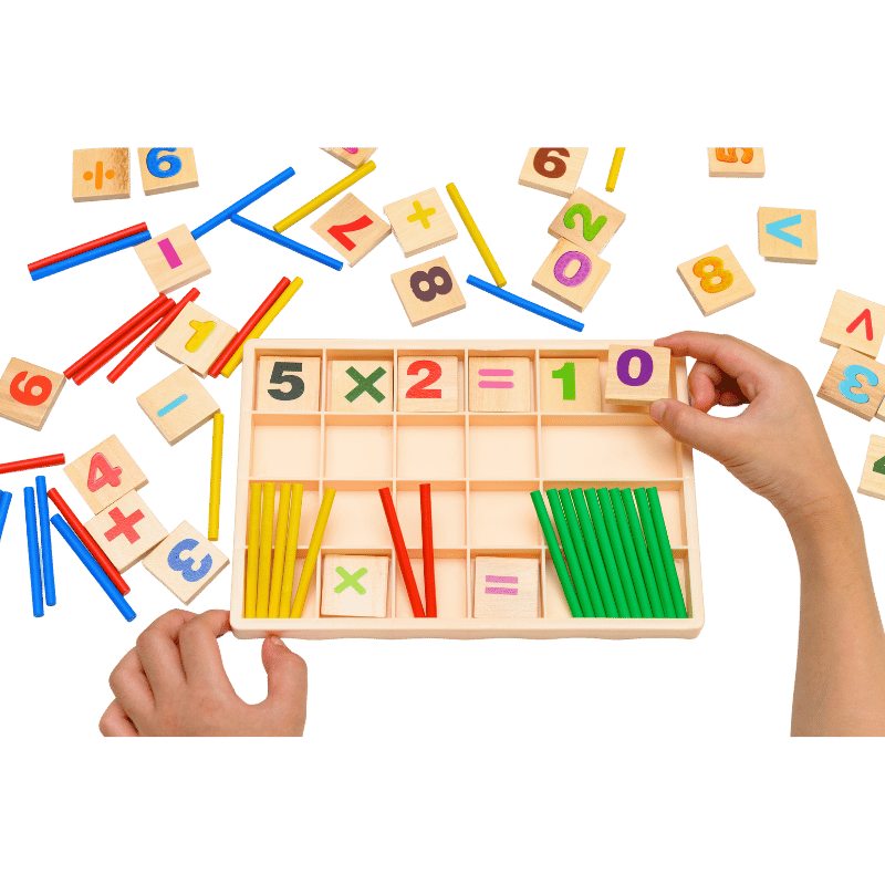 Hands arrange number tiles on a colorful math board showing 5×2=10, with scattered wooden number blocks on a green background.