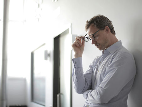 stressed looking man leans against wall, taking off his glasses.