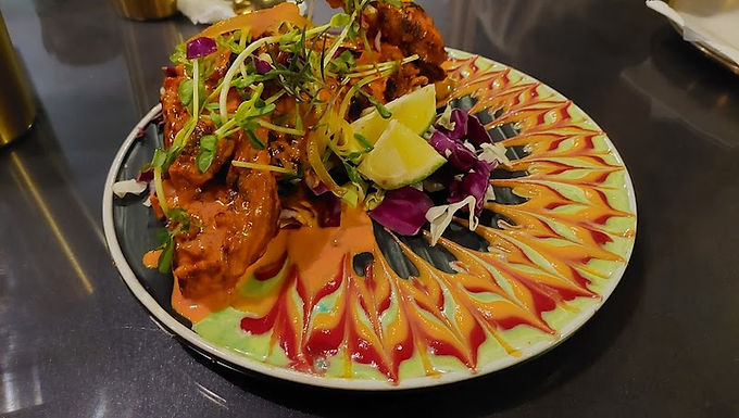 坦都里烤雞（Tandoori Chicken，又稱印度烤雞）是一道源自印度次大陸、極受歡迎的南亞經典料理，其製法是將帶骨雞肉浸泡在優格和被稱為「坦都里瑪薩拉」（Tandoori Masala）的濃郁綜合香料混合物中，利用優格的酸性使肉質軟嫩，並以辣椒粉等香料賦予雞肉鮮豔的紅或橘色。醃製後的雞肉會串上長籤，放入傳統的 坦都里爐（Tandoor，一種高溫黏土烤爐）中炙烤，爐內的高熱能迅速鎖住肉汁，並產生獨特的煙燻焦香和微焦的外皮，使其口感異常鮮嫩多汁，通常會搭配檸檬汁一同享用。

Tandoori chicken is a celebrated South Asian dish where bone-in chicken is marinated in a mixture of yogurt and tandoori masala—a robust blend of spices including ginger, garlic, cayenne, and garam masala—to tenderize the meat and impart a deep red hue. This richly flavored chicken is traditionally cooked on long skewers inside a tandoor, a high-heat, cylindrical clay oven, where the intense heat seals in the juices and creates a characteristic smoky aroma and slightly charred exterior, resulting in an exceptionally tender and flavorful dish, often enjoyed with lemon wedges.