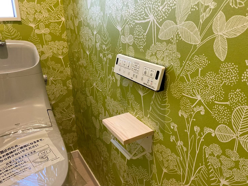 緑の花柄壁紙のトイレ内。左にINAXの便器、壁に操作パネルと折りたたみ棚があり、便座に注意書きの日本語が見える。