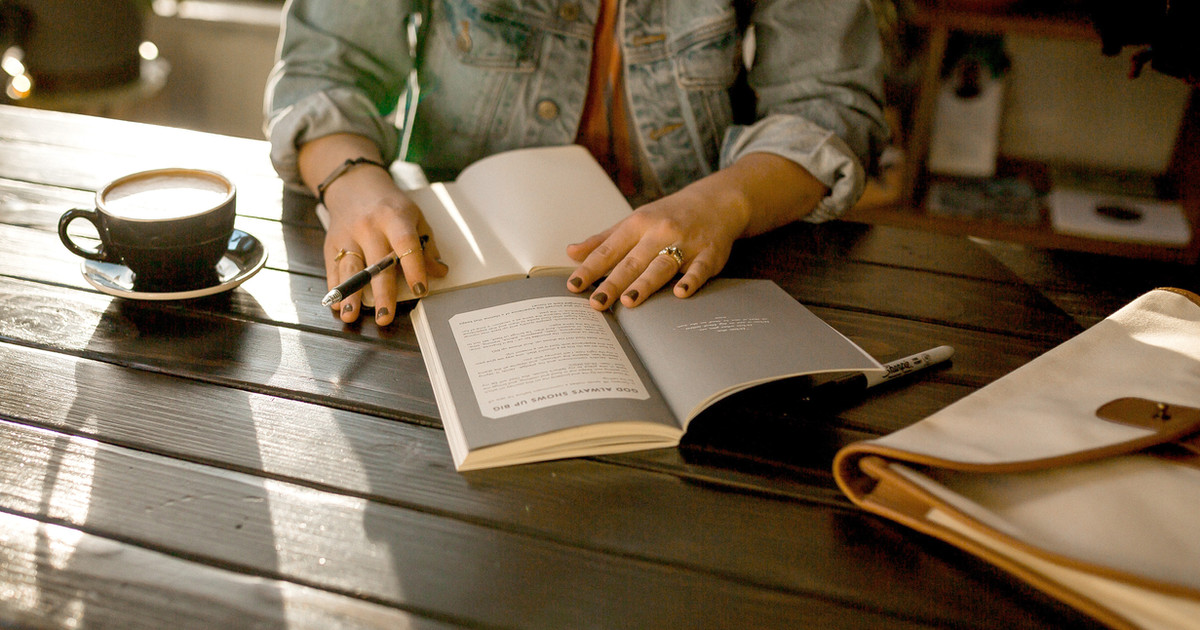 A person writing in a small notebook at a kitchen table, with a cup of tea nearby, in soft morning light.