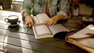 Person in a denim jacket reads a book at a wooden table with a cup of coffee and bag nearby. Warm sunlight creates a cozy atmosphere.