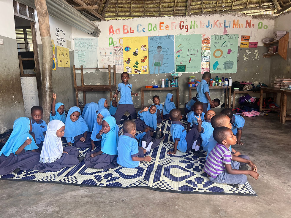 Children in blue uniforms and hijabs sit on a patterned mat in a classroom with colorful educational posters and the alphabet on the wall.