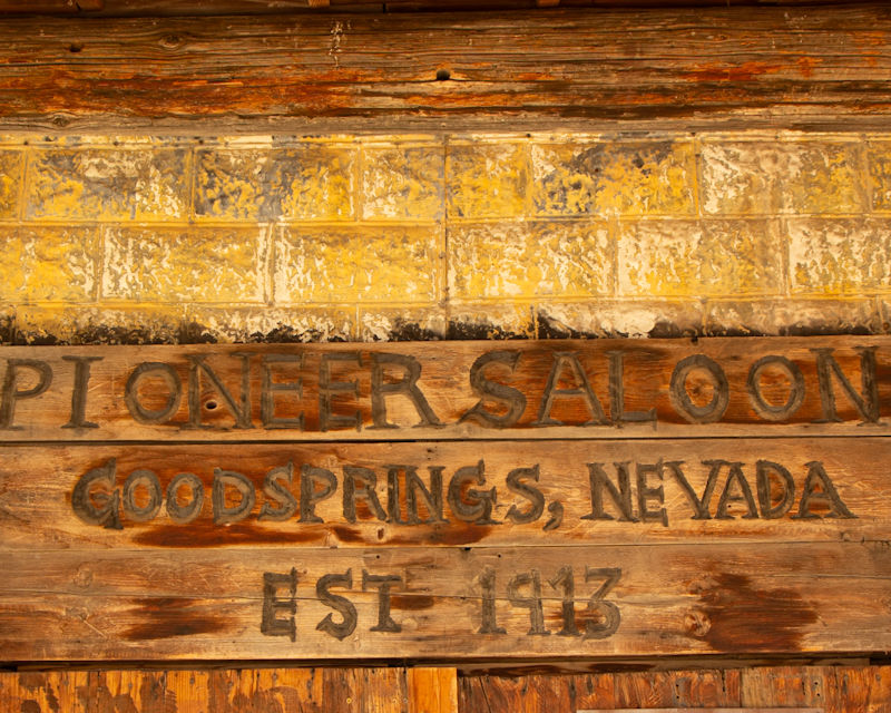 Weathered sign reading "Pioneer Saloon, Goodsprings, Nevada, Est. 1913" on rustic wood with yellow brick background, rustic and nostalgic mood.