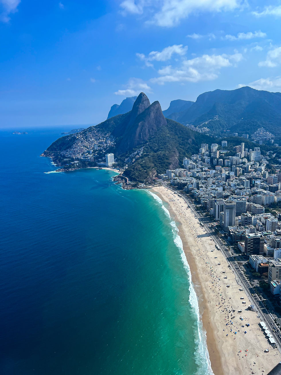 Aerial view of Rio de Janeiro's coastline with turquoise sea, sandy beach, city buildings, and mountains under a bright blue sky.