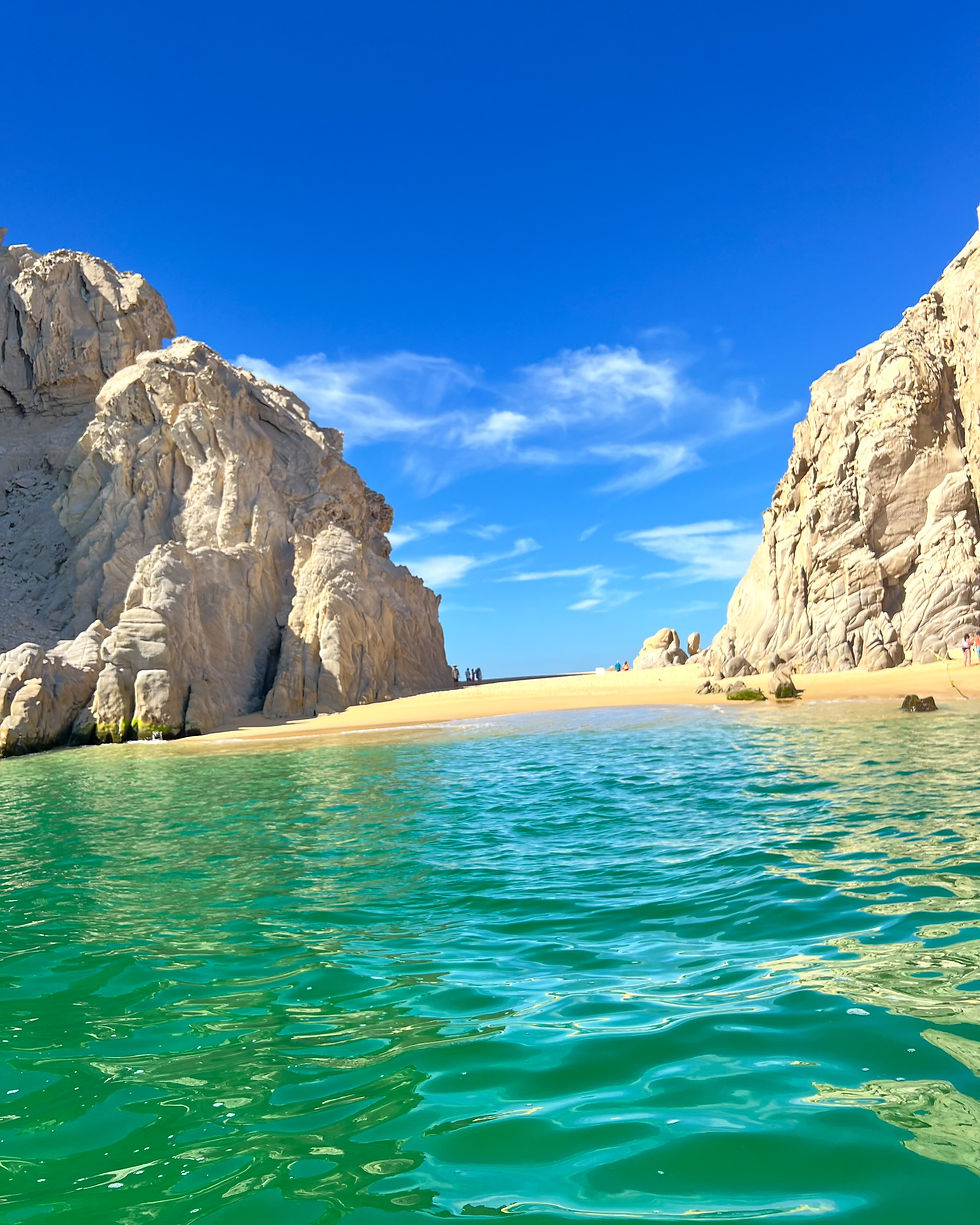 Turquoise water flows between towering beige cliffs under a clear blue sky, leading to a sandy beach in the distance.