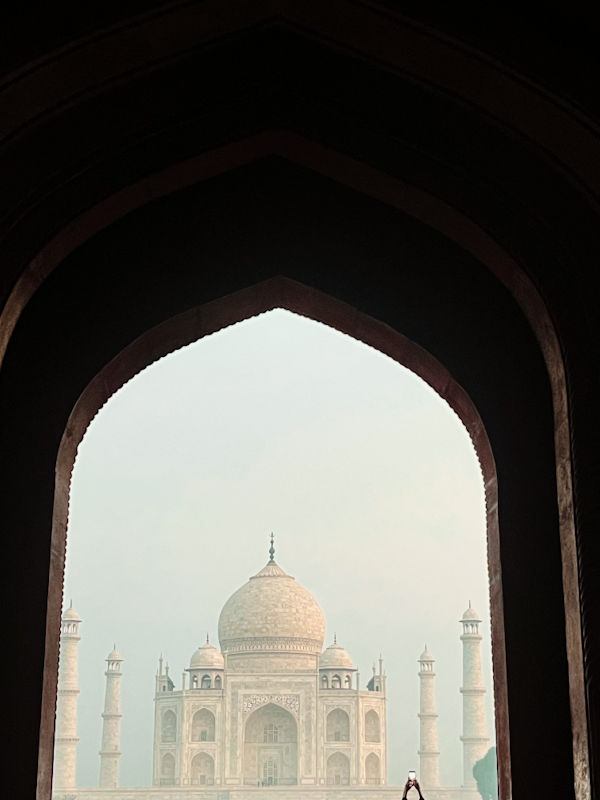 Taj Mahal framed by a dark archway, standing against a hazy sky. A person is visible in front, offering a serene and iconic view.