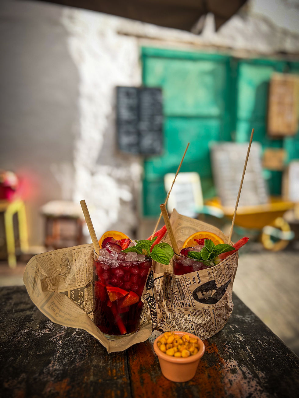 Two iced drinks wrapped in newspaper, garnished with orange slices and basil, on a rustic table with a cup of nuts. Green door background.