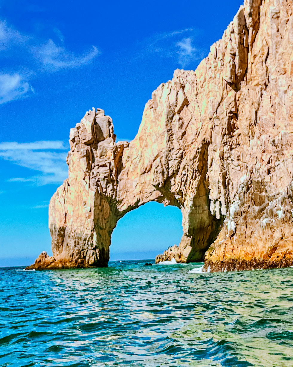 Natural rock arch over turquoise water under a clear blue sky. Rugged cliffs rise dramatically, with sunlight casting shadows. Calm, scenic mood.