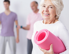 Older lady smiling in white Tshirt holding a rolled up pink yoga mat with blurred other people in the background doing exercise