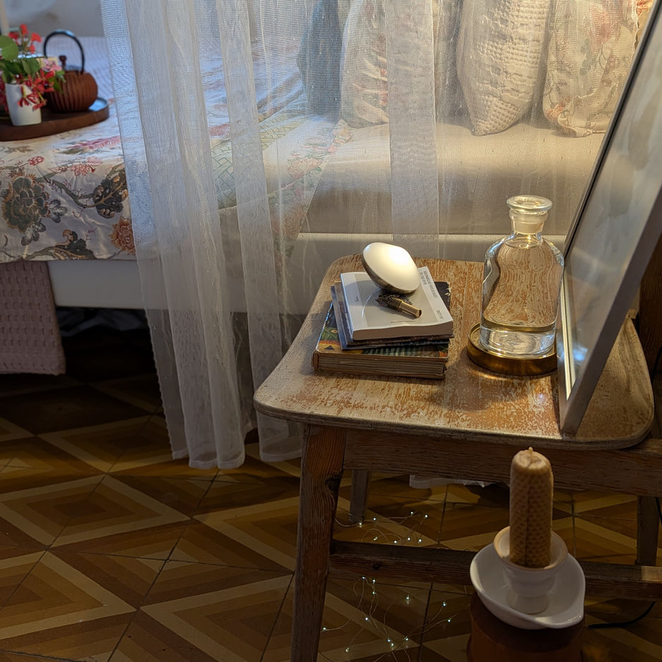 Mesa de noche de madera con libros, botella de agua y vasos, lamparas de noche, y la cama de fondo con las texturas y transparencias de las telas.
