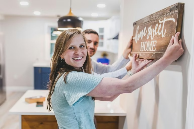 A smiling couple hangs a wooden “home sweet home” sign on a wall in their bright
kitchen.