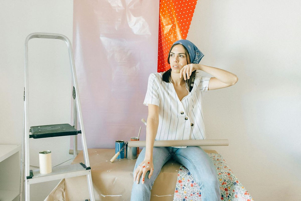 Woman in striped shirt and jeans sits on a floral-covered surface, holding a cardboard tube. Paint cans and ladder nearby, wall art behind.
