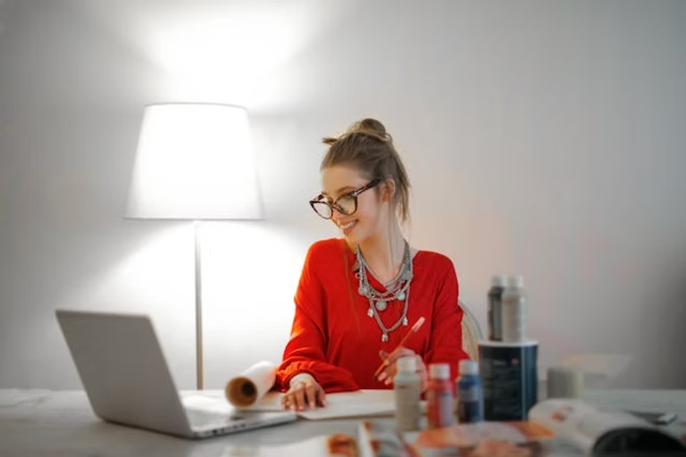 Woman in a red shirt sitting at a table with a laptop, paint bottles, and an open magazine. She's smiling, under a lit lamp, in a cozy room.
