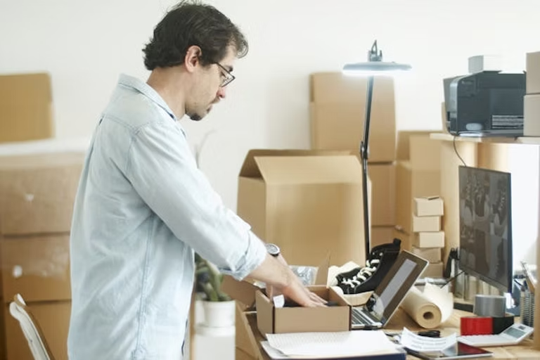 Man packing boxes in a room filled with cardboard boxes, using a laptop. Sneakers, rolls of paper, and a lamp are visible. Bright setting.