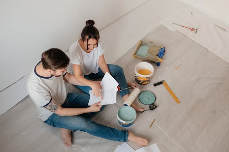 A couple sitting on a floor covered with plastic, reading instructions near open paint cans and rollers