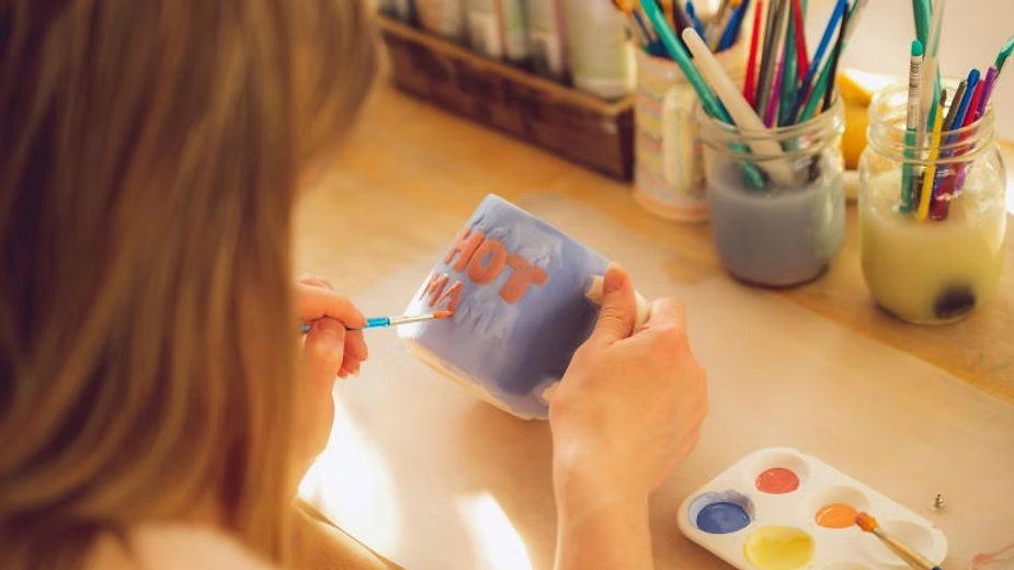 Woman painting a ceramic mug with orange letters at a desk filled with jars of brushes and paints.