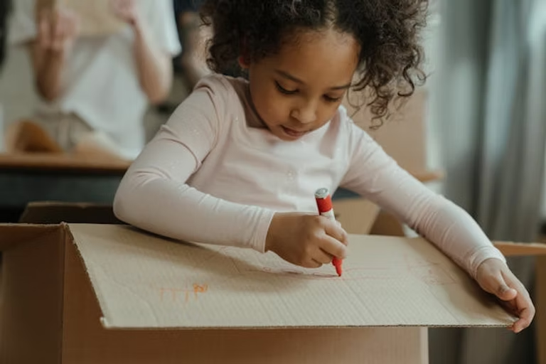 Young girl draws on a cardboard box with a red marker, focused and calm. Wearing a white top, background shows a blurred person.