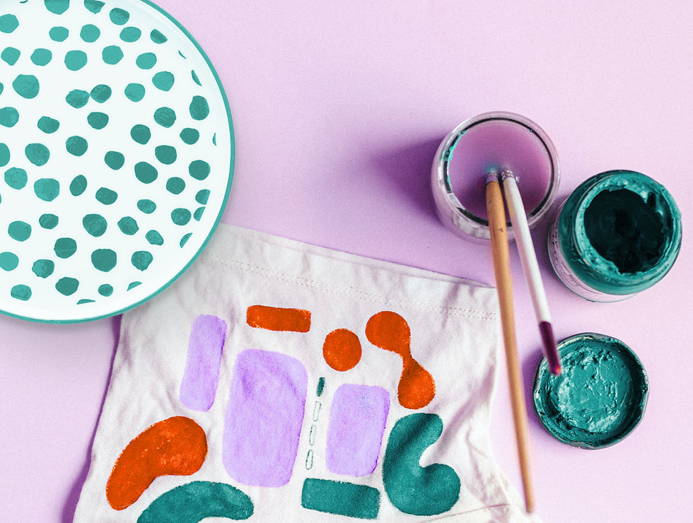 Art supplies on a pink surface, featuring colorful painted fabrics, a dot-patterned plate, paint jars, and brushes, creating a playful mood.
